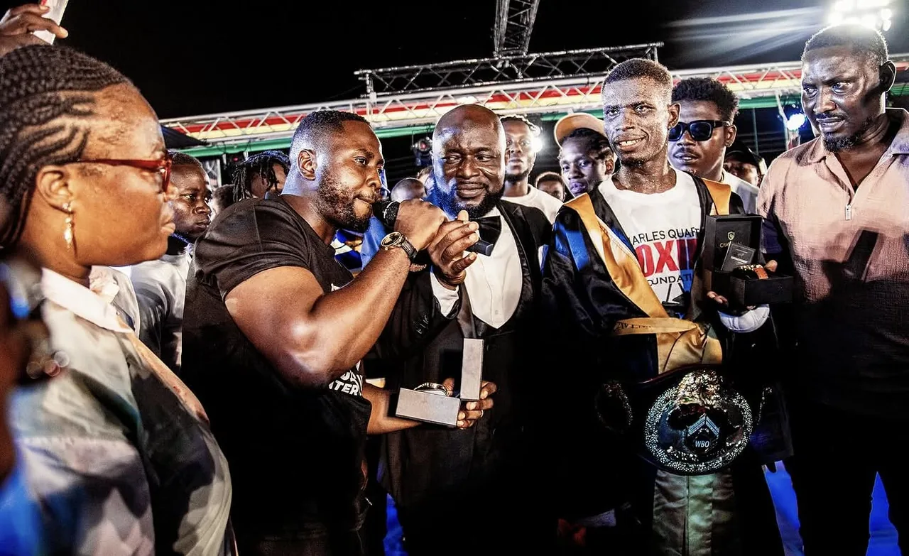 Edwin Elikem Amenyeawu speaking ringside as a Ghanaian boxer holds his WBO Africa title belt